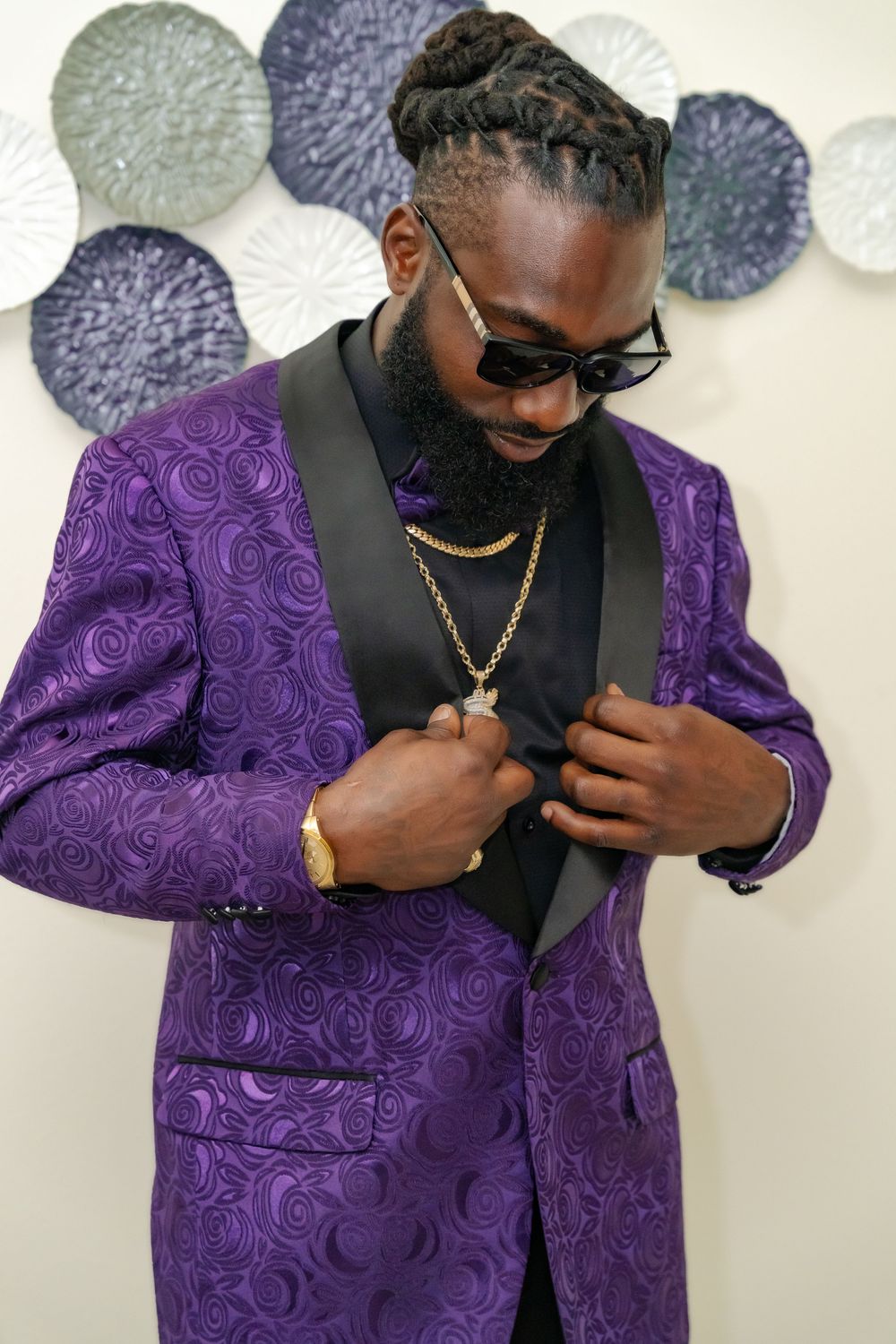 Groom adjusting jacket wearing purple paisley tuxedo against wall with decorative circular elements.