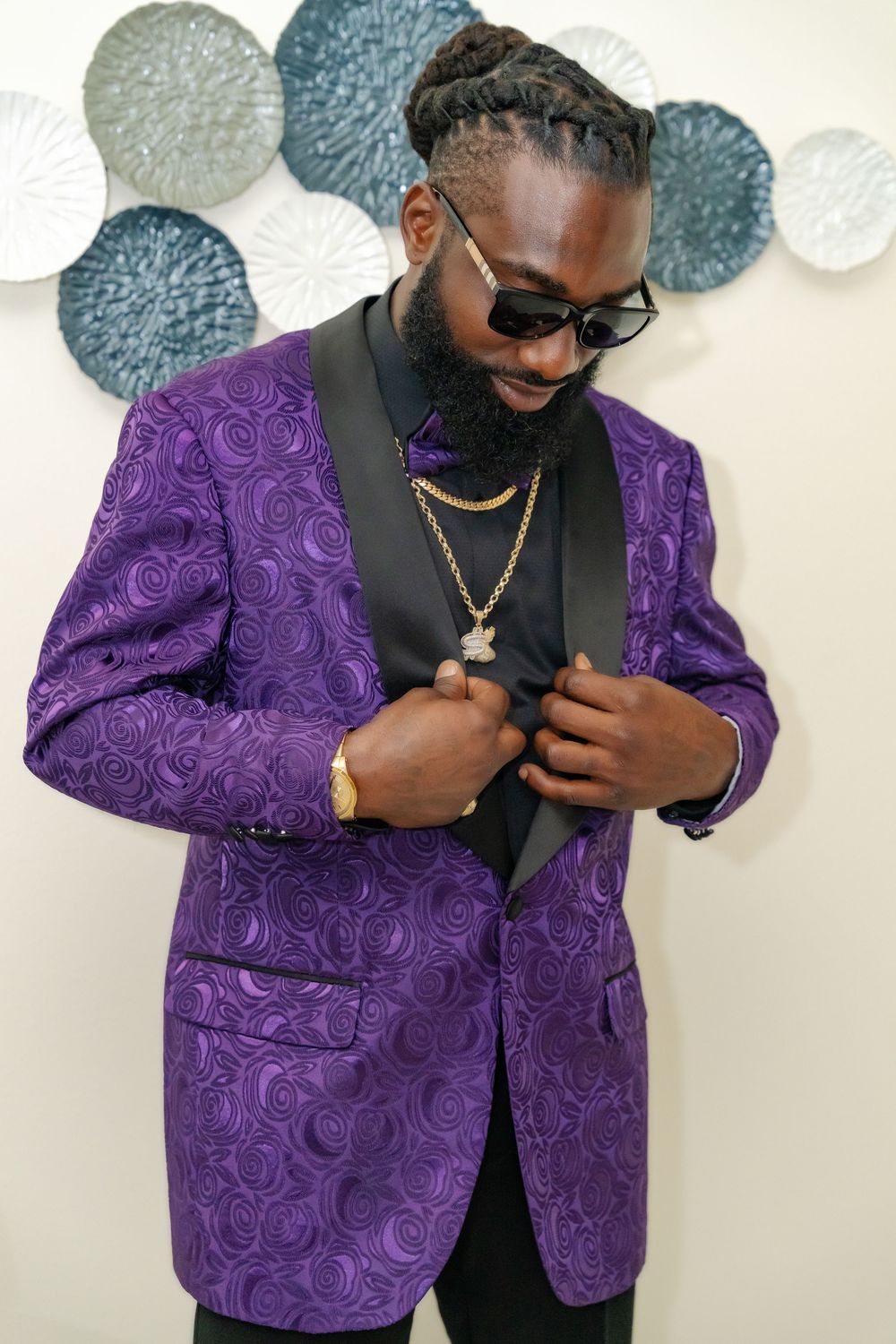 Side view of groom in purple patterned suit jacket and sunglasses against decorated wall.