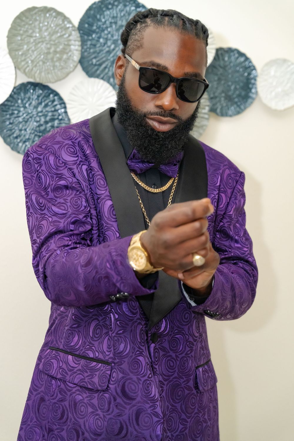 Groom checking gold watch while wearing purple paisley suit and sunglasses.