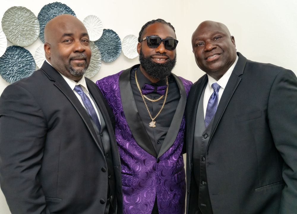 Men in black suits and one in a purple vest stand together against a white wall with decorative accents.
