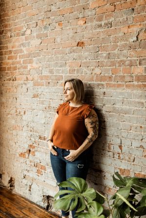 A person in an orange top poses against a rustic brick wall with indoor plants.