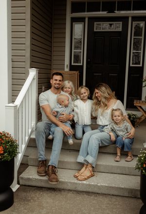 A family sits together on the front porch steps of their home with white siding and a black door.