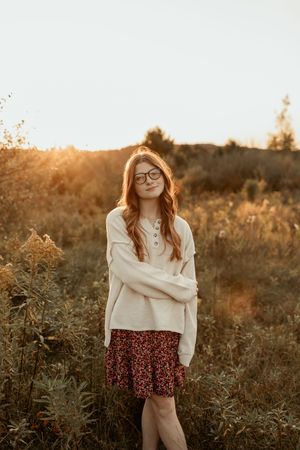 A figure in a cream sweater and red floral skirt stands in a wild meadow at golden hour.