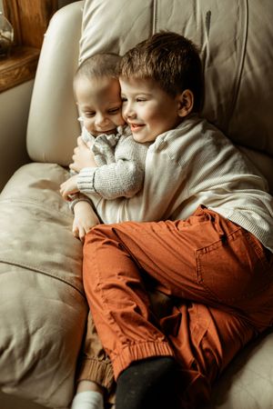 Two children cuddling on a beige leather couch, one wearing orange pants and one in a light colored outfit.