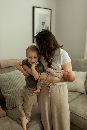 Woman in white shirt and beige pants embraces children on a gray couch in a cozy living room setting.