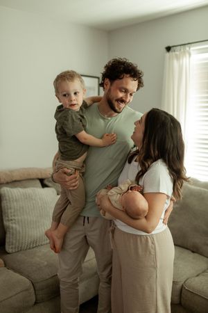 A candid moment showing a couple in neutral-toned clothing playing with a toddler on a gray sofa in a bright living room.