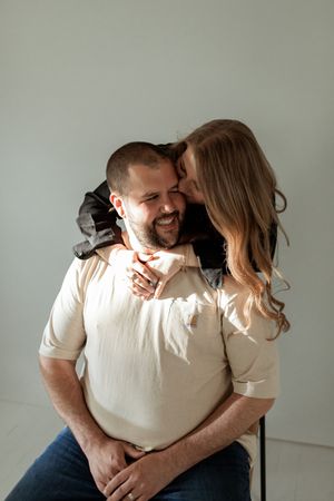 Two people in white shirts share intimate laughter and embraces while sitting against a plain wall.