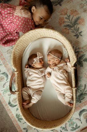 Two newborn babies wearing matching pink outfits lie sleeping together in a round wicker bassinet.
