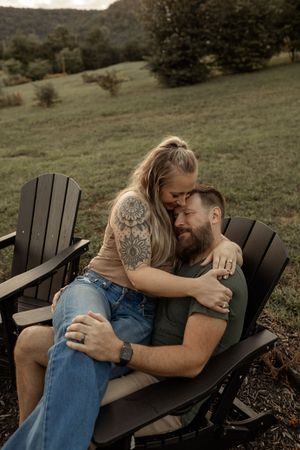 A couple shares an intimate embrace while sitting on an Adirondack chair outdoors on a hillside.