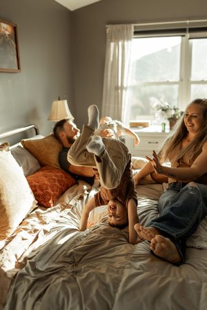 A family relaxes together on a bed on a sunny afternoon, sharing playful moments in natural light streaming through windows.