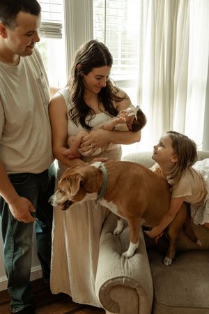 Family in white loungewear poses lovingly with their brown dog on a gray couch in a sun-lit living room.