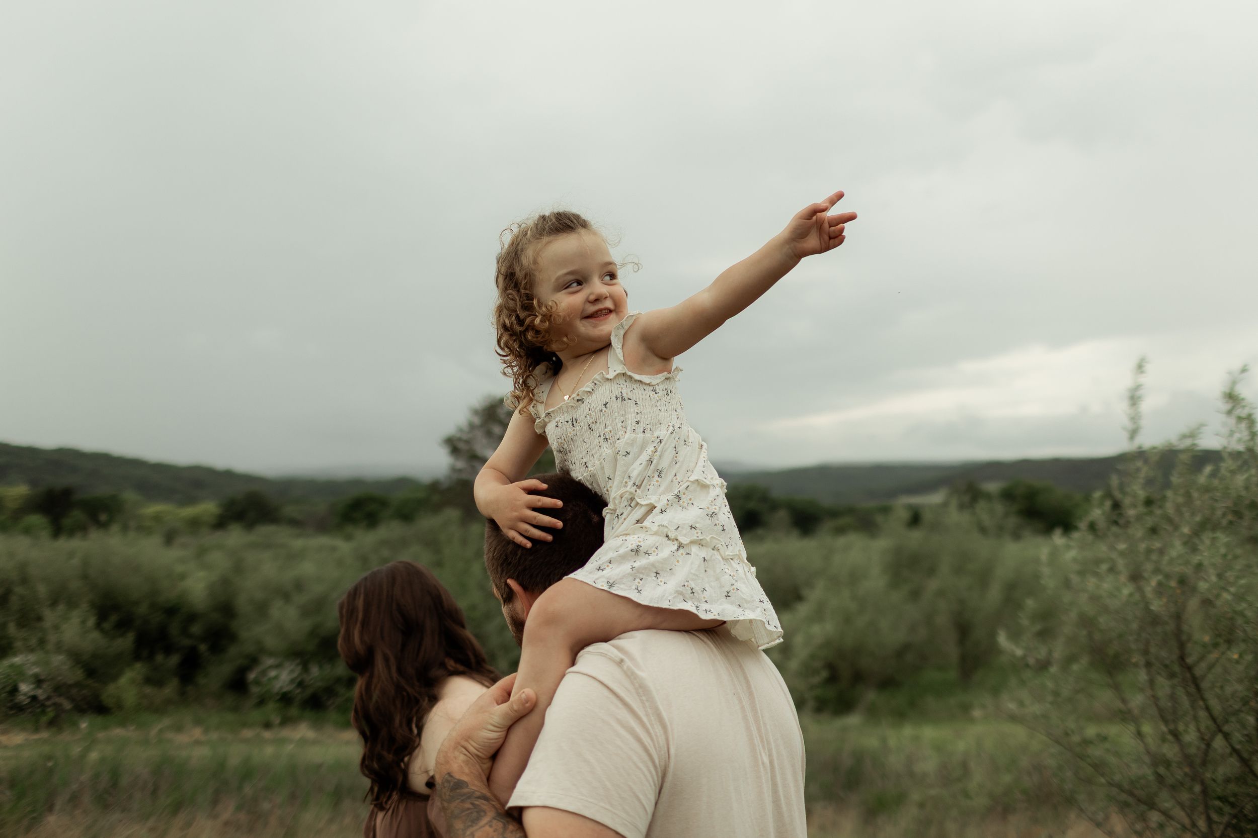 A parent in beige dress lifts child wearing white dress joyfully in an outdoor setting with green fields and cloudy sky.