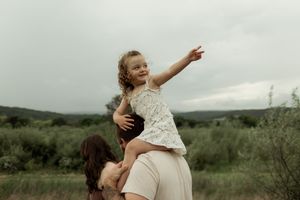 A parent in beige dress lifts child wearing white dress joyfully in an outdoor setting with green fields and cloudy sky.