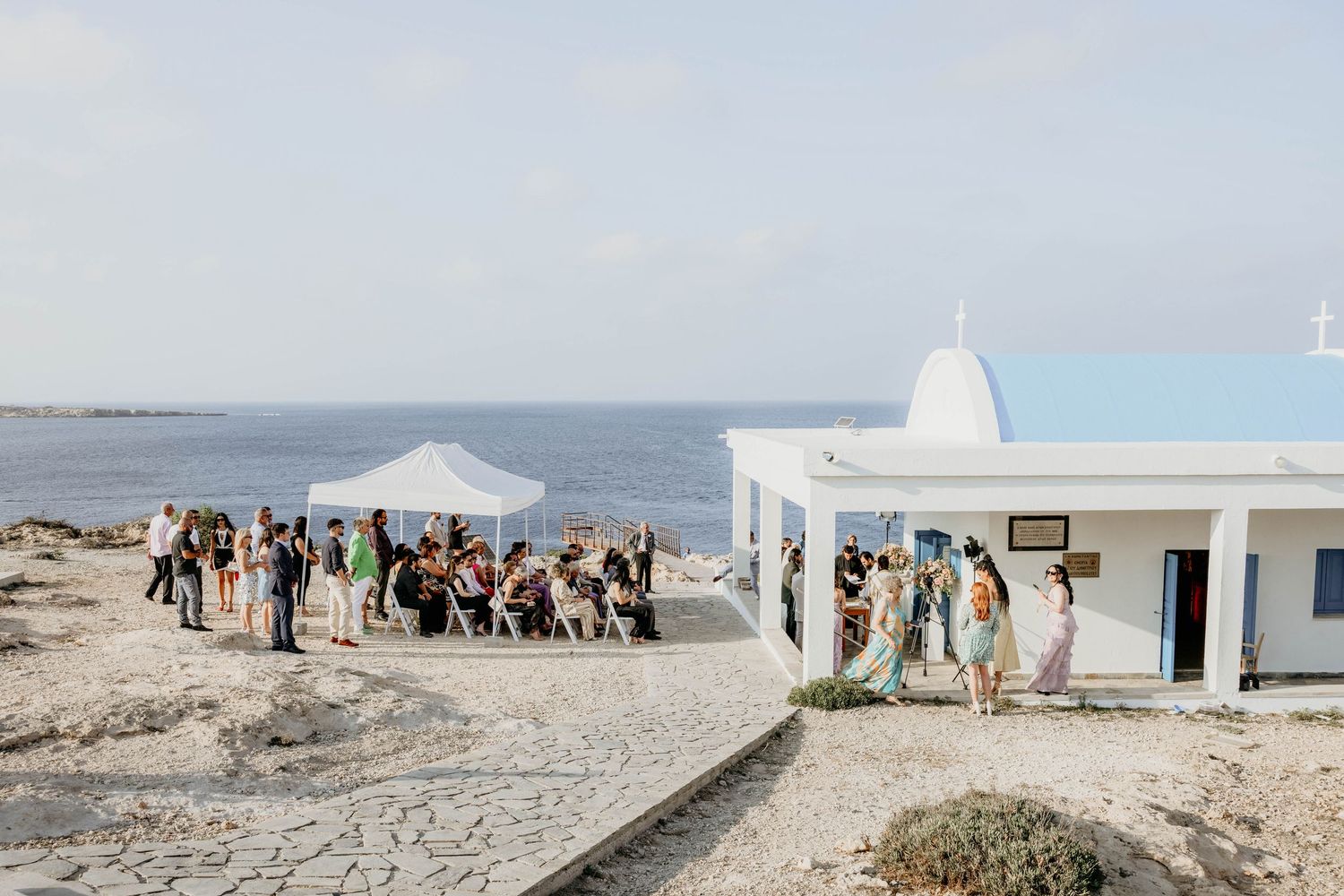 Several people walk along a scenic white coastal path near the Mediterranean Sea with traditional architecture visible.