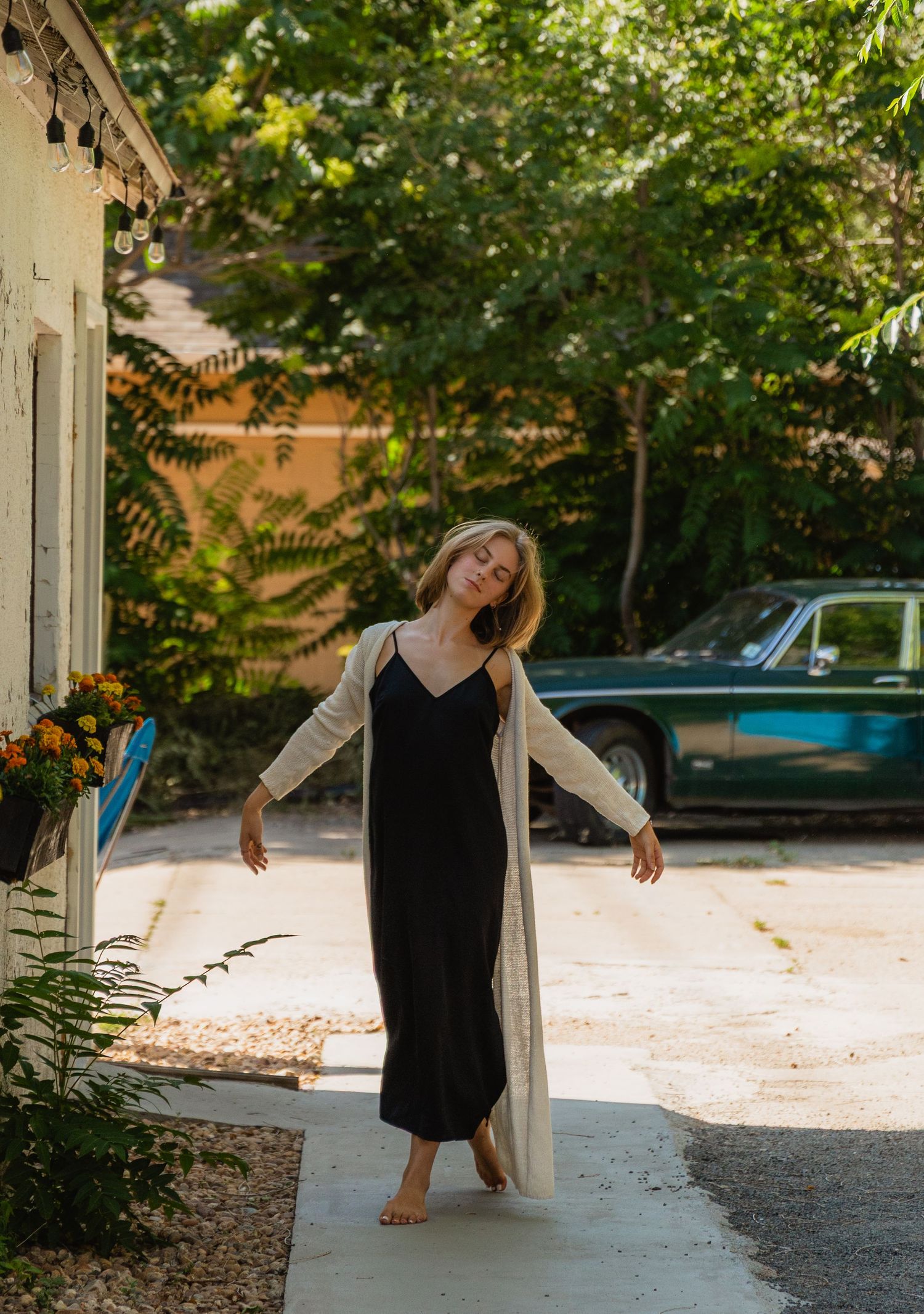 A person in a black slip dress and long beige cardigan stands barefoot on a sidewalk in dappled sunlight.