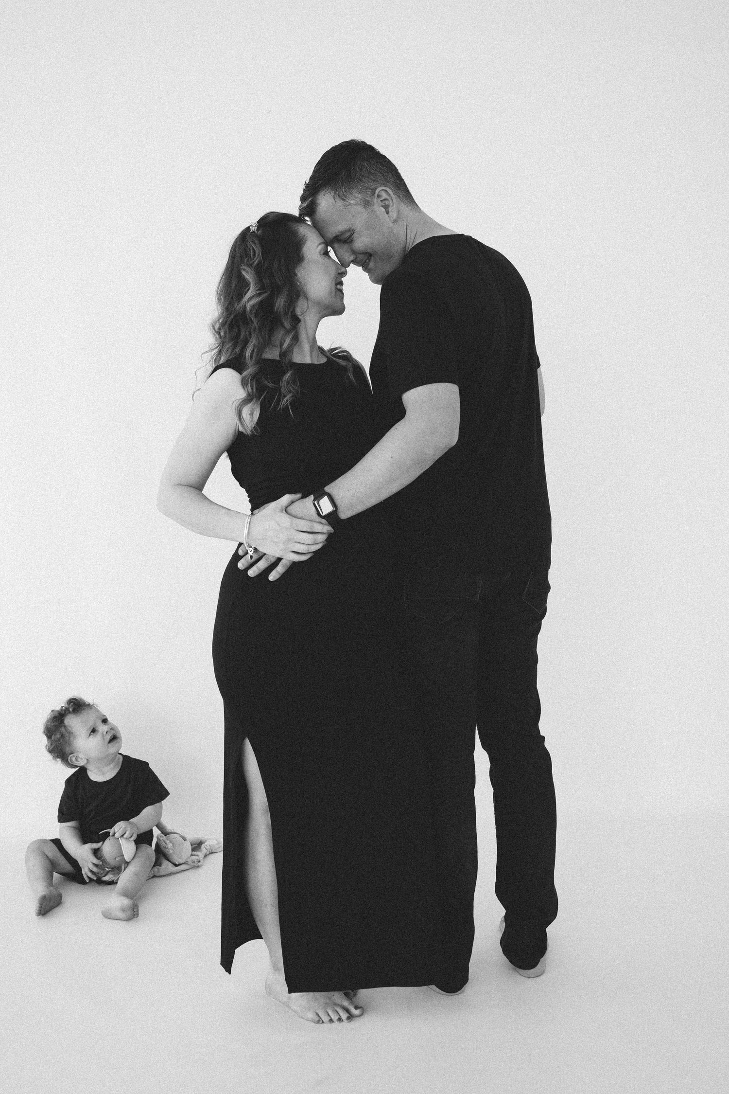Black and white portrait of a romantic couple in formal wear embracing while a small child sits on the floor nearby.