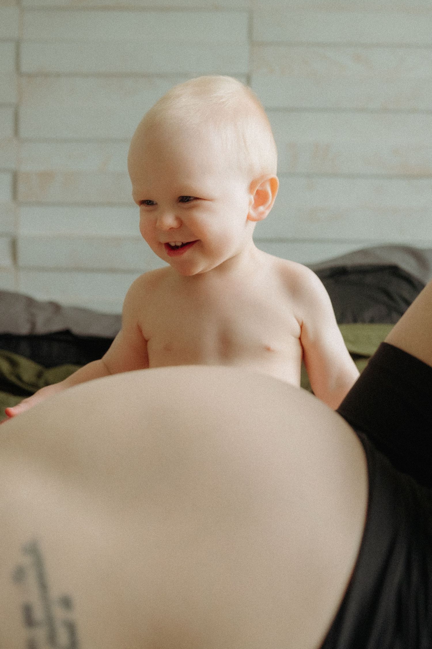 A happy baby with light hair smiles while leaning against a pregnancy bump in a warm indoor setting.
