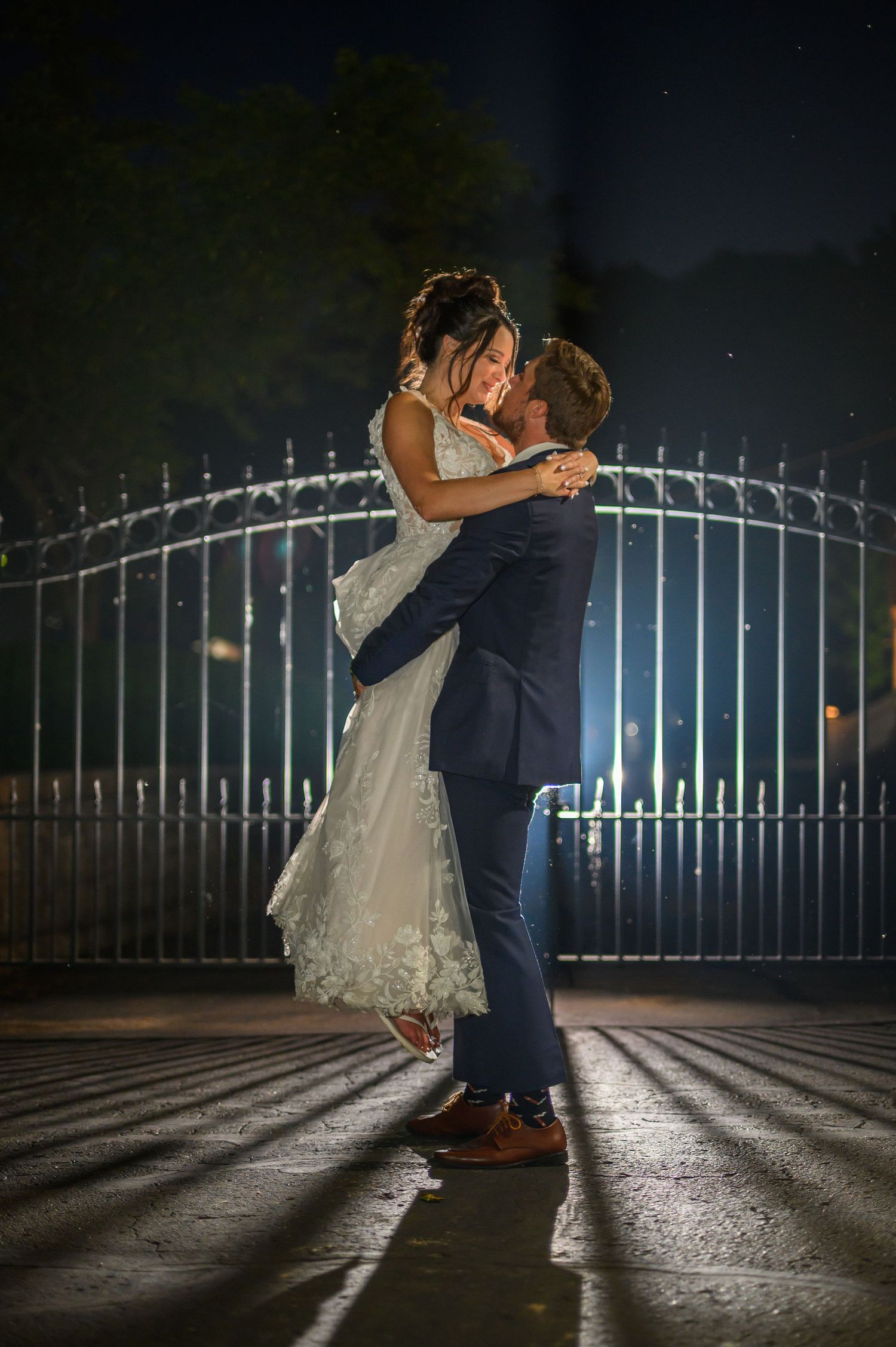 A romantic nighttime wedding photo shows a couple dancing in front of an illuminated iron gate casting dramatic shadows.