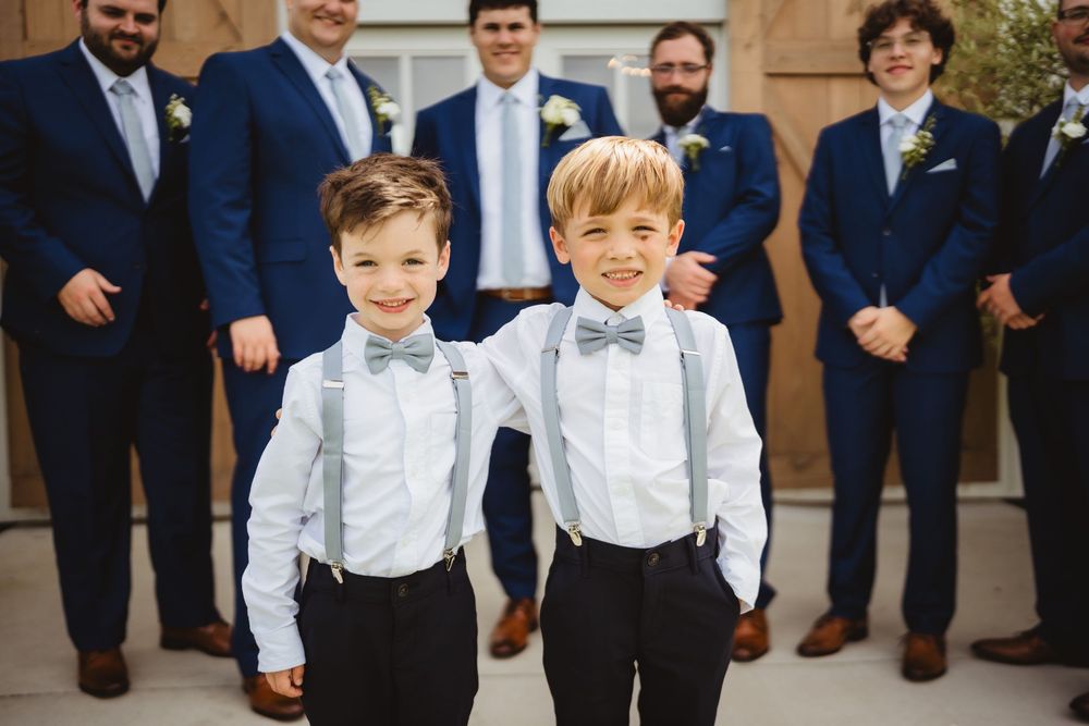 Two young ring bearers in white shirts with bow ties and suspenders stand in front of groomsmen in navy suits at a wedding.