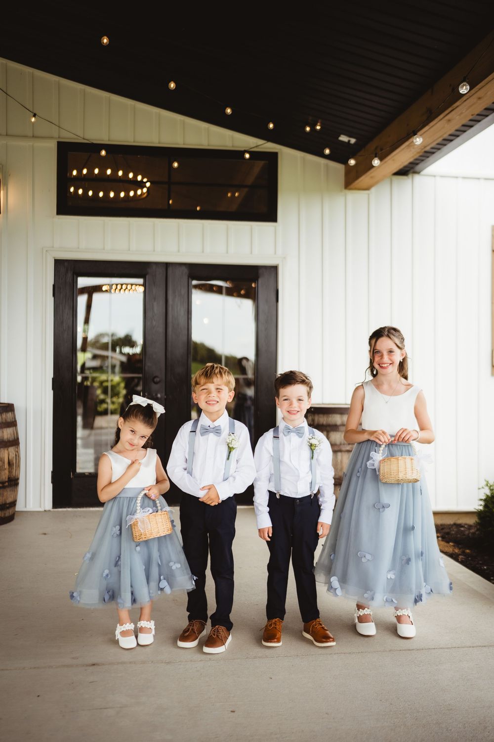 Wedding party children in coordinating blue and white formal attire stand in front of modern barn venue entrance.