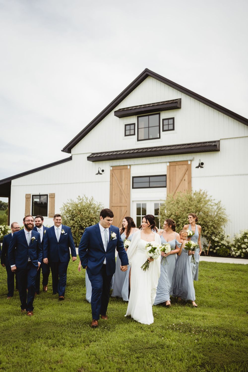 Wedding party walks together on grass in front of a modern white barn with wooden barn doors and window frames.