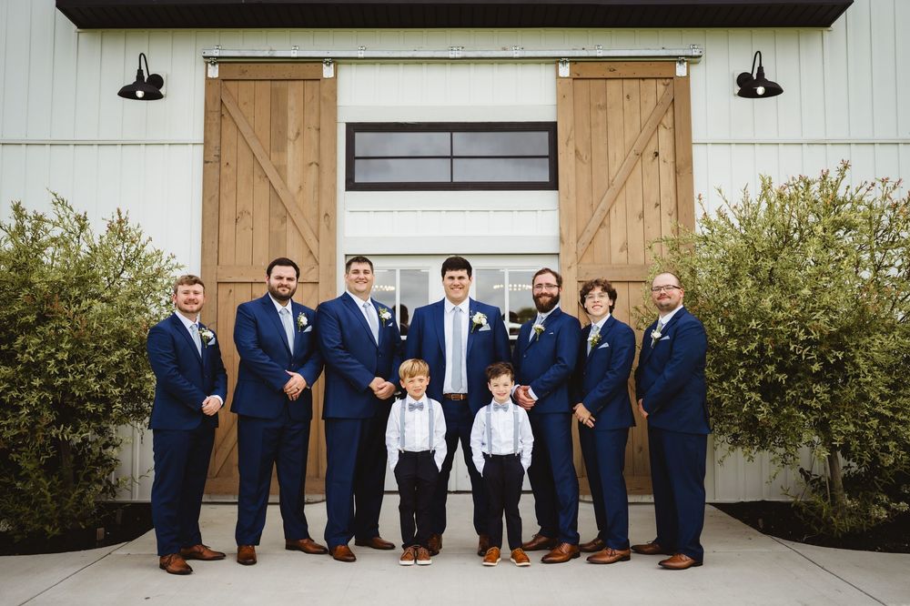 Wedding party members in navy suits stand together against a rustic outdoor backdrop during formal portraits.