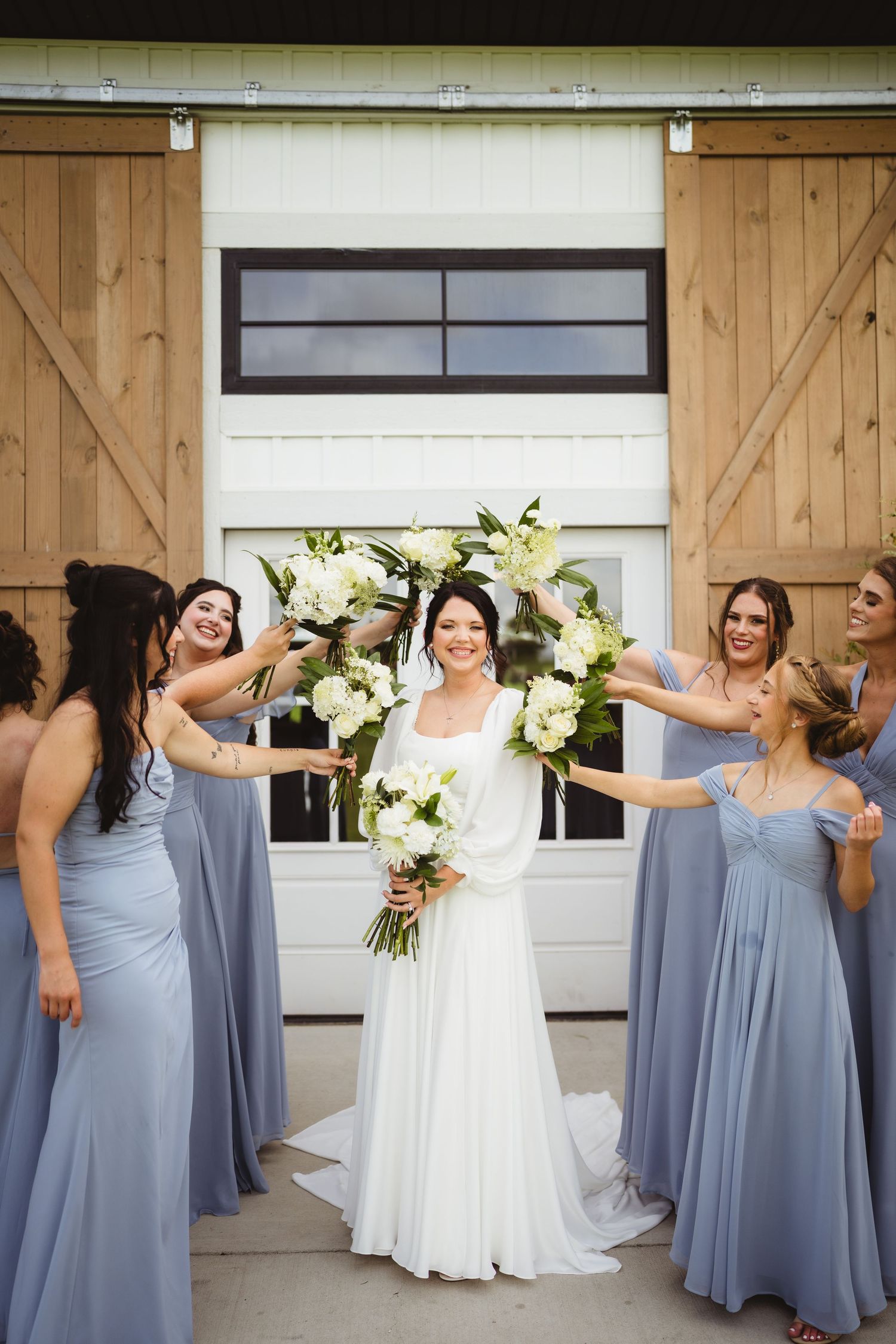 A bridal party in flowing dusty blue bridesmaid dresses and a white wedding gown pose in front of a barn door.