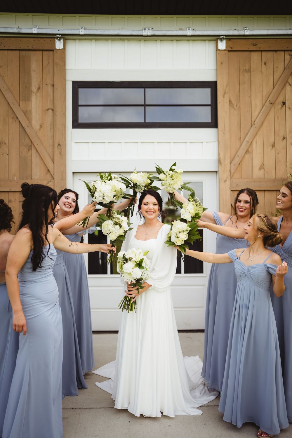 A bridal party in flowing dusty blue bridesmaid dresses and a white wedding gown pose in front of a barn door.