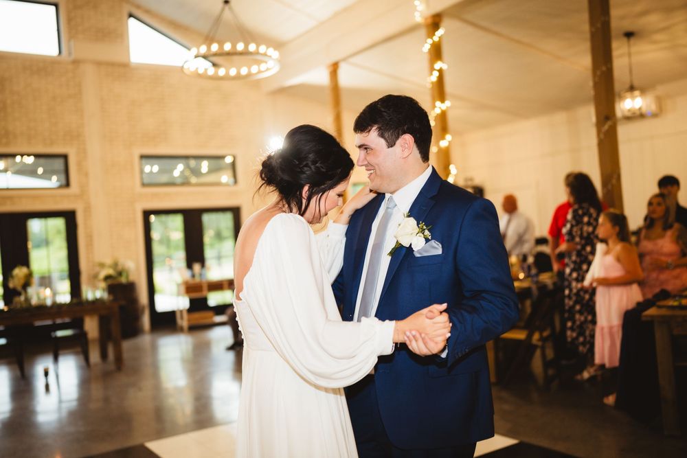 A bride and groom share their first dance at an elegant reception venue with warm lighting and large windows.