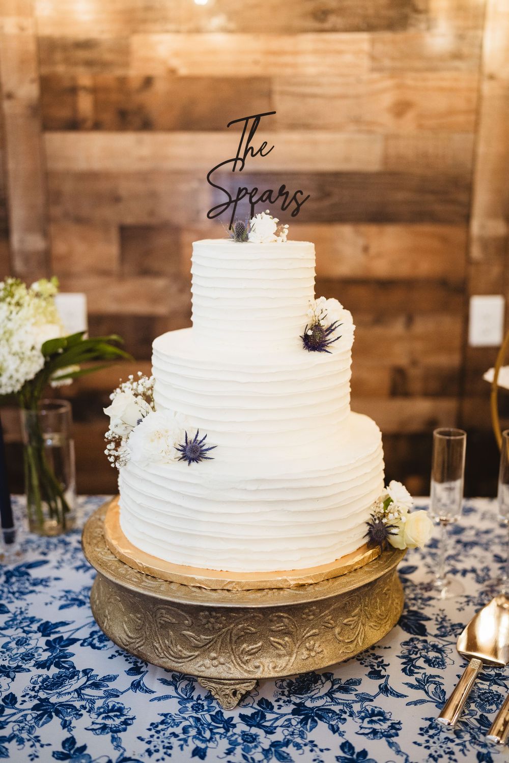Simple white tiered wedding cake decorated with delicate lines and small purple thistle flowers on a gold stand with blue tablecloth.