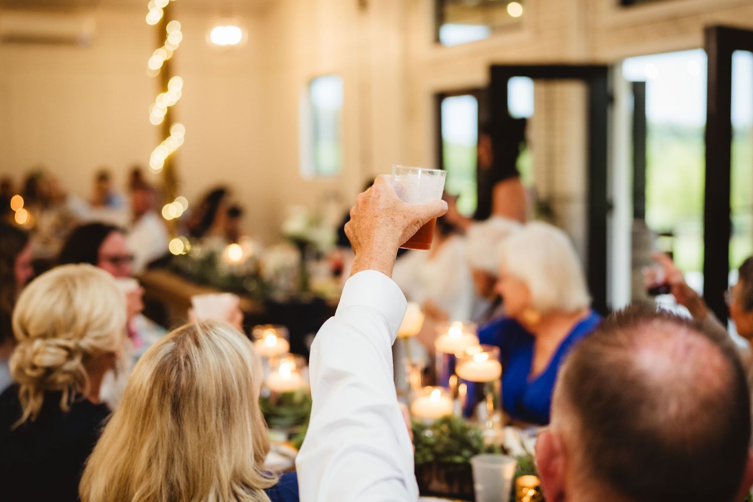 A person raises a glass in celebration during a festive dinner reception with lights and other guests visible in background.