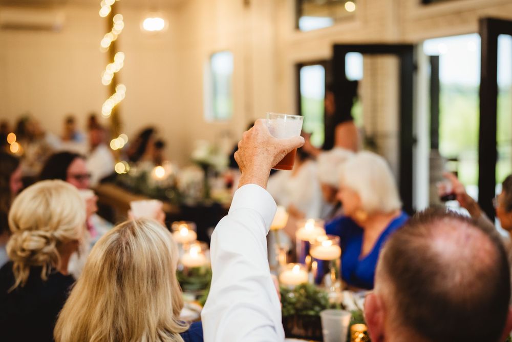 A person raises a glass in celebration during a festive dinner reception with lights and other guests visible in background.