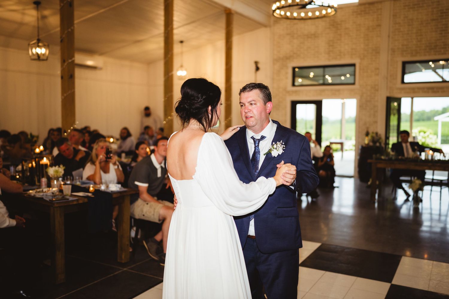 A couple shares their first dance in an elegant reception venue with rustic wooden tables and chandeliers.