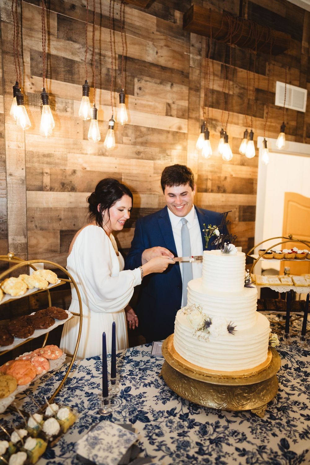A couple cuts into their elegant white wedding cake at a rustic venue with wooden walls and hanging lights.