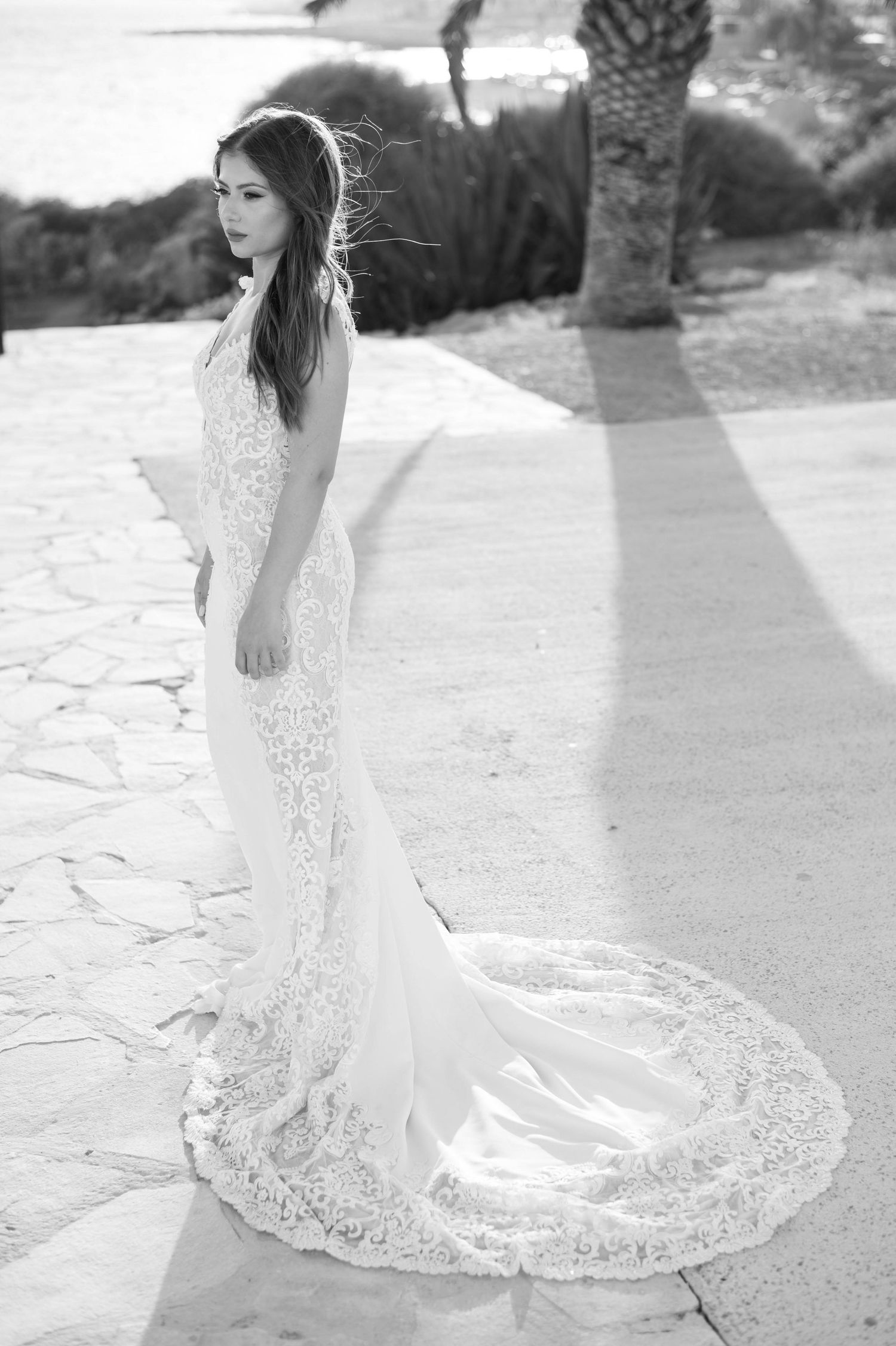 A bride in an elegant white lace wedding dress with a flowing train poses gracefully outdoors in black and white photography.