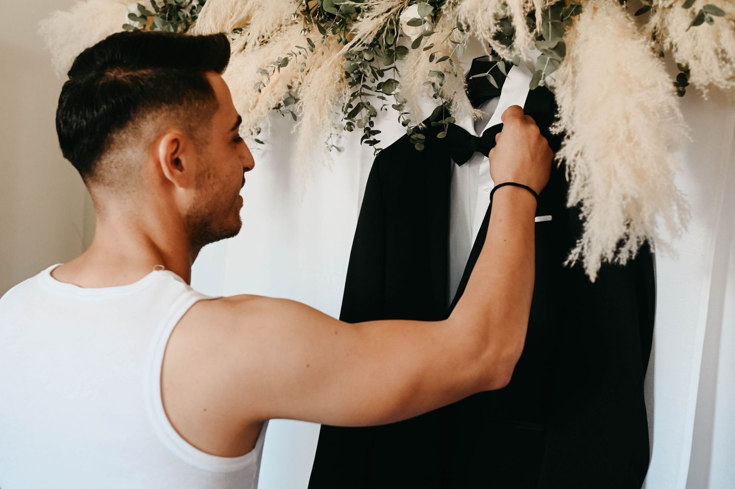 A person in a white tank top arranges dried pampas grass flowers against a dark wall for decoration.