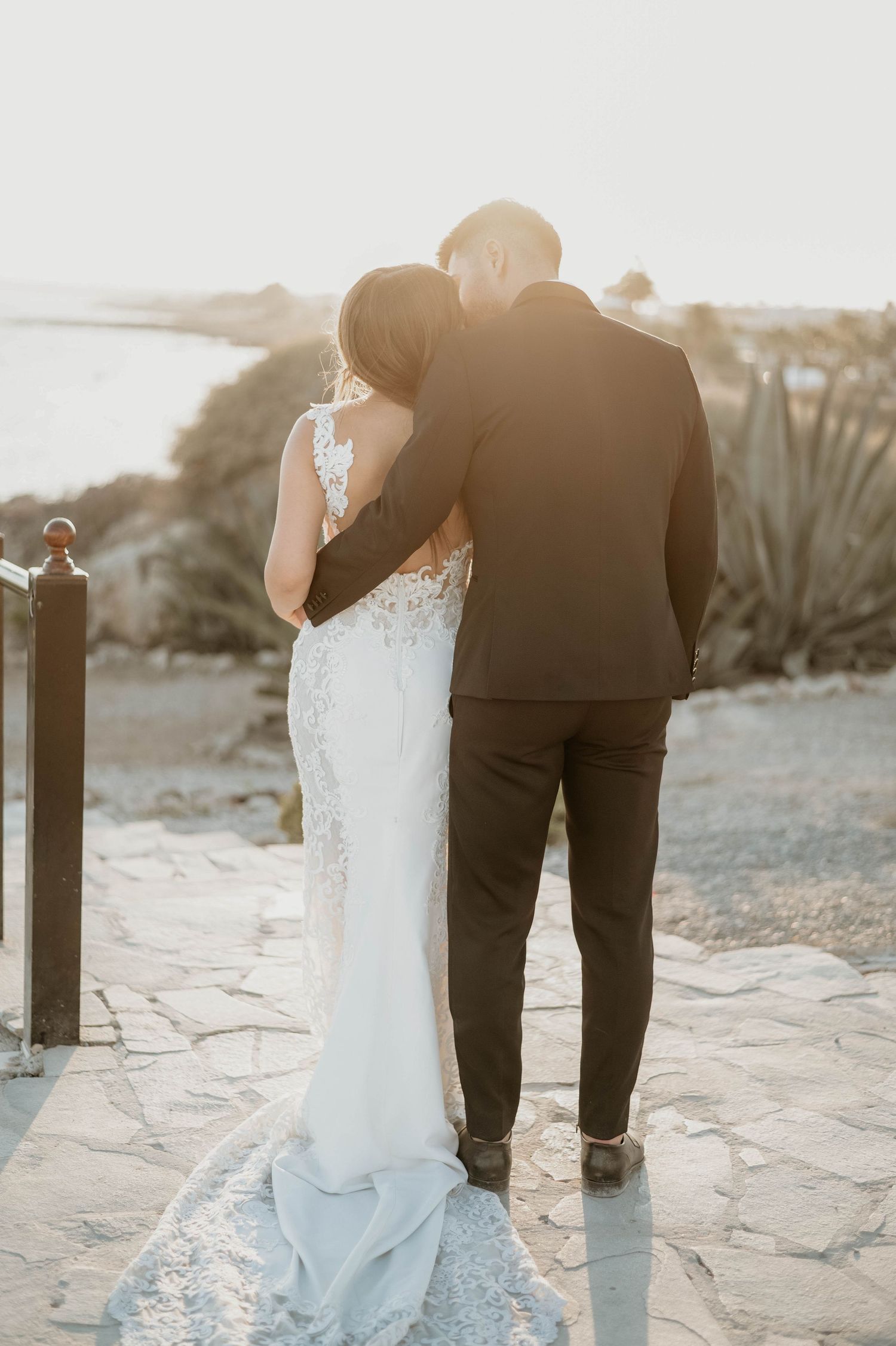 Romantic wedding photo of couple embracing on stone patio overlooking scenic outdoor view at sunset.
