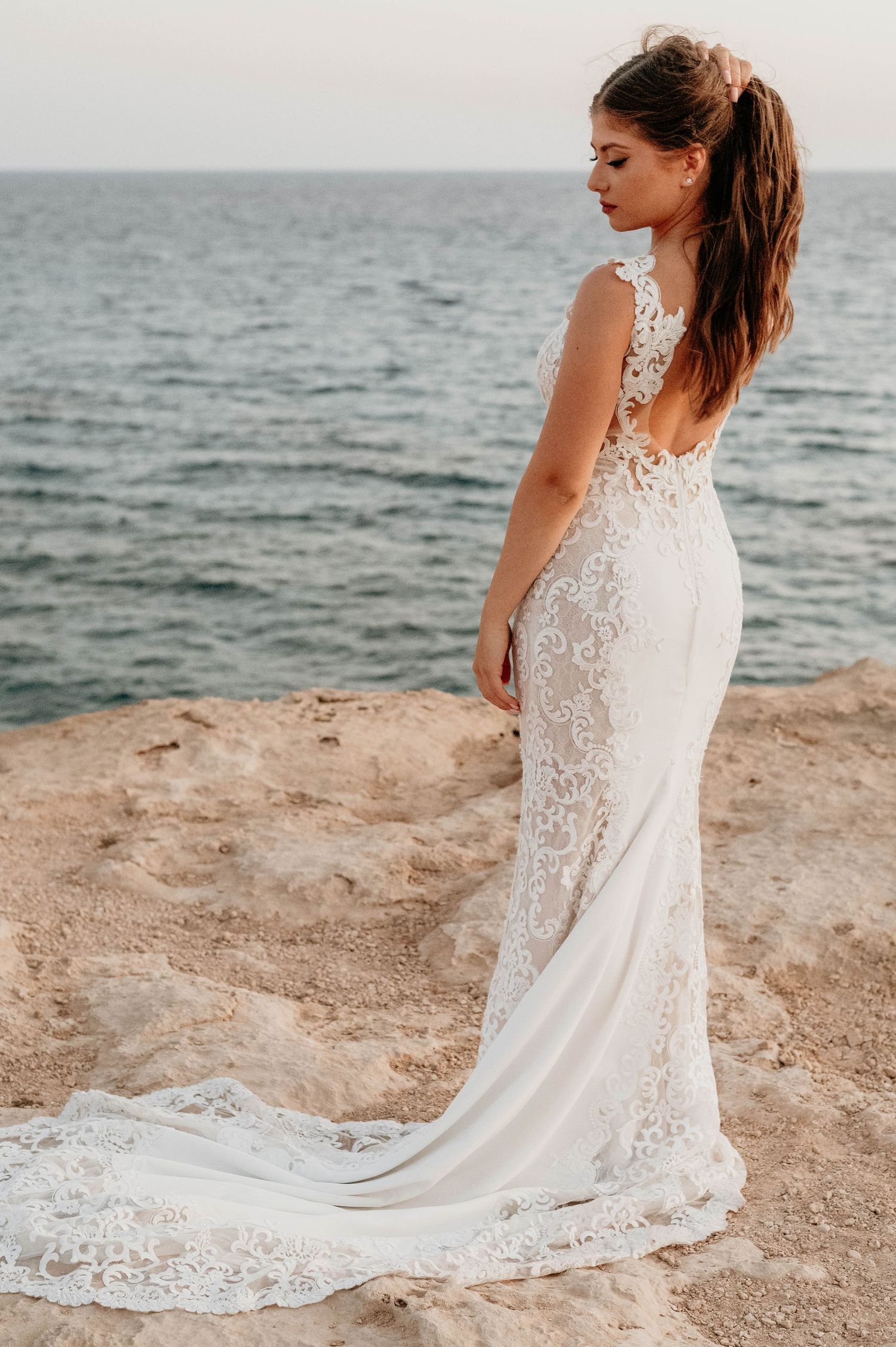A bride in a flowing white lace wedding dress stands on rocky cliffs overlooking the ocean at sunset.
