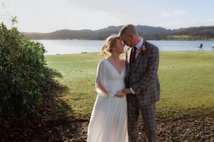 A couple shares an intimate moment on a grassy waterfront with mountains and a lake in the background.