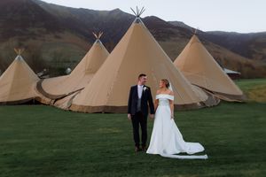 Wedding couple stands in front of three tipi tents at a rustic outdoor venue with mountains in background.