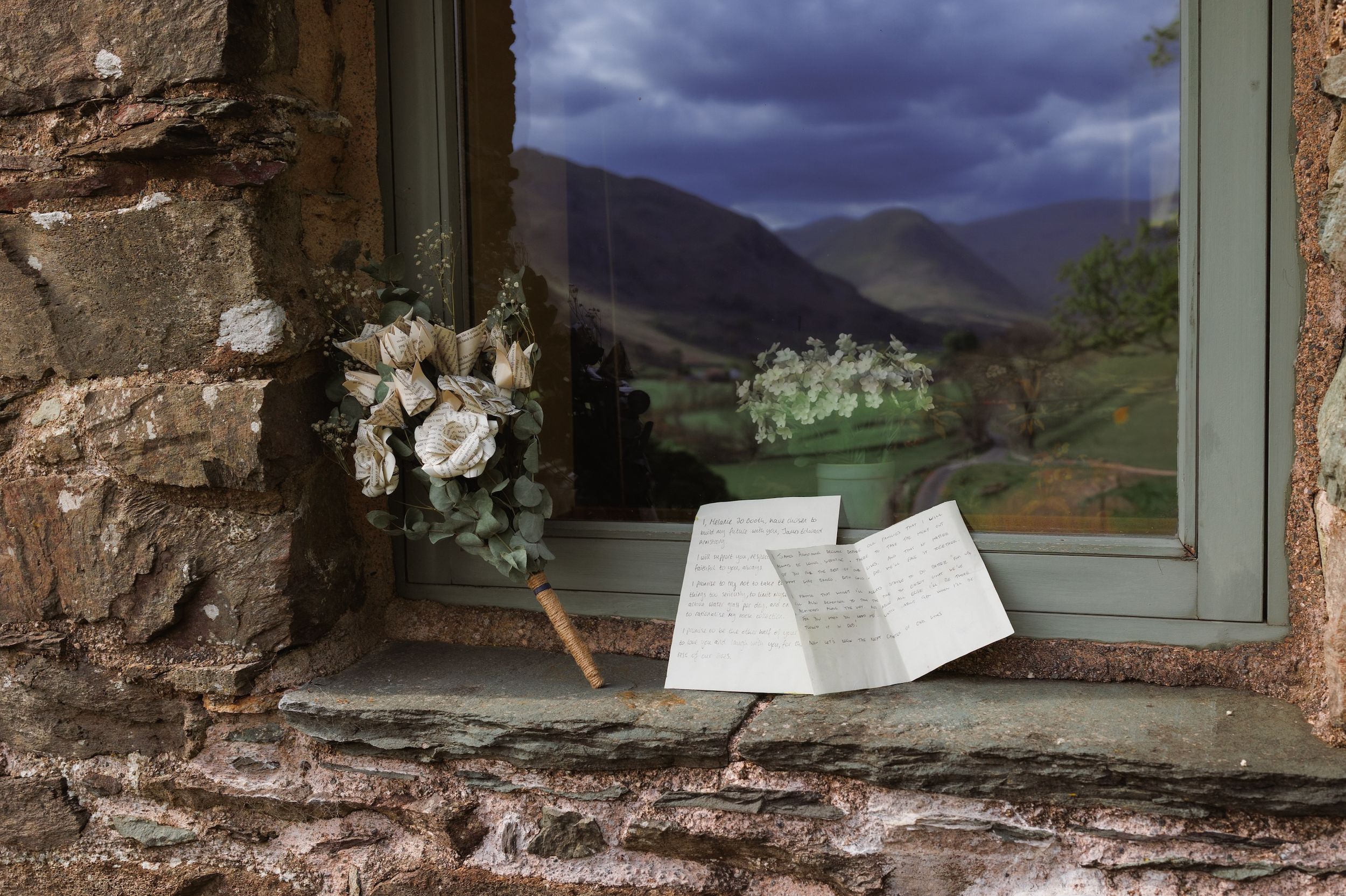 A handwritten letter and white flowers rest on a rustic stone windowsill overlooking mountain scenery.