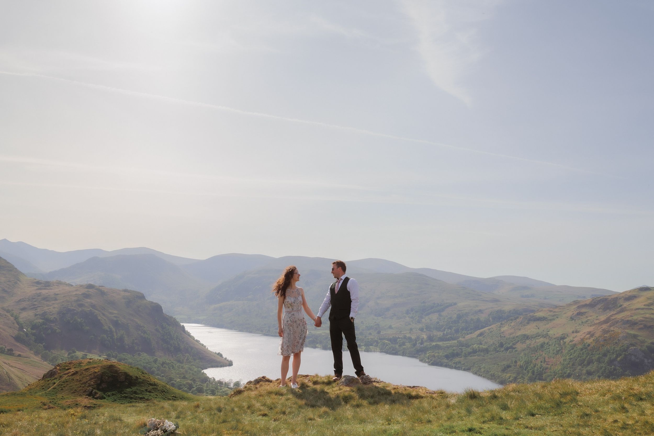 Two figures stand atop a scenic overlook with mountain ranges and a winding lake visible in the background.