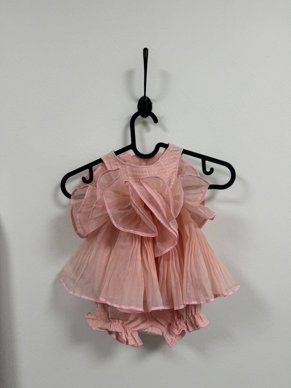 Pink ruffled baby dress with matching bloomers hanging on black plastic hangers against white wall.