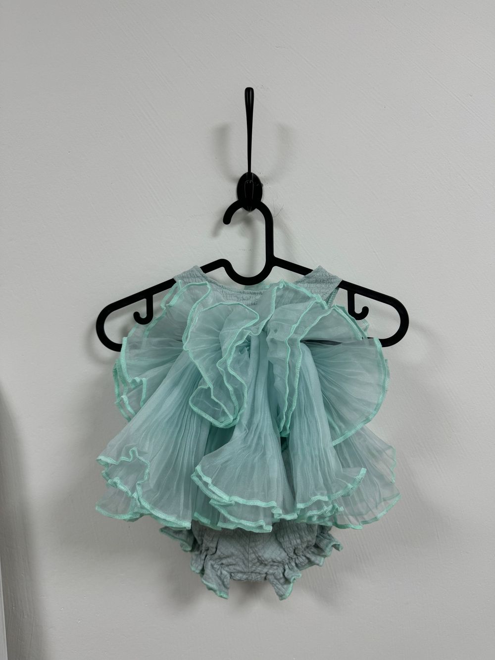 A delicate mint green ruffled tutu dress with matching bloomers hangs on a black plastic hanger against a white wall.