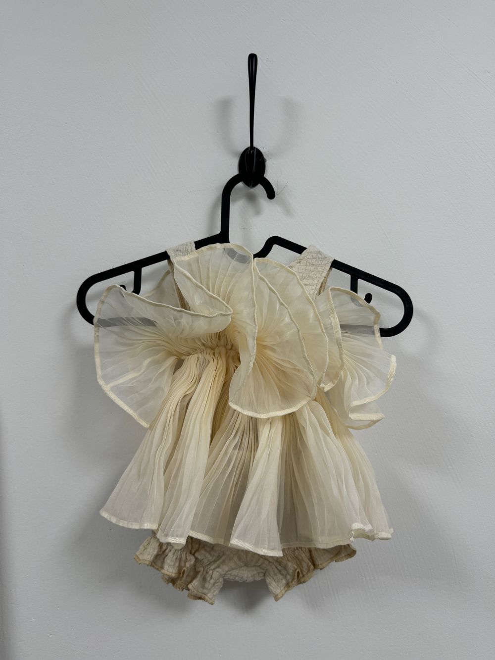 A delicate cream-colored vintage ruffle dress with pleated details hangs on a black plastic hanger against a white wall.