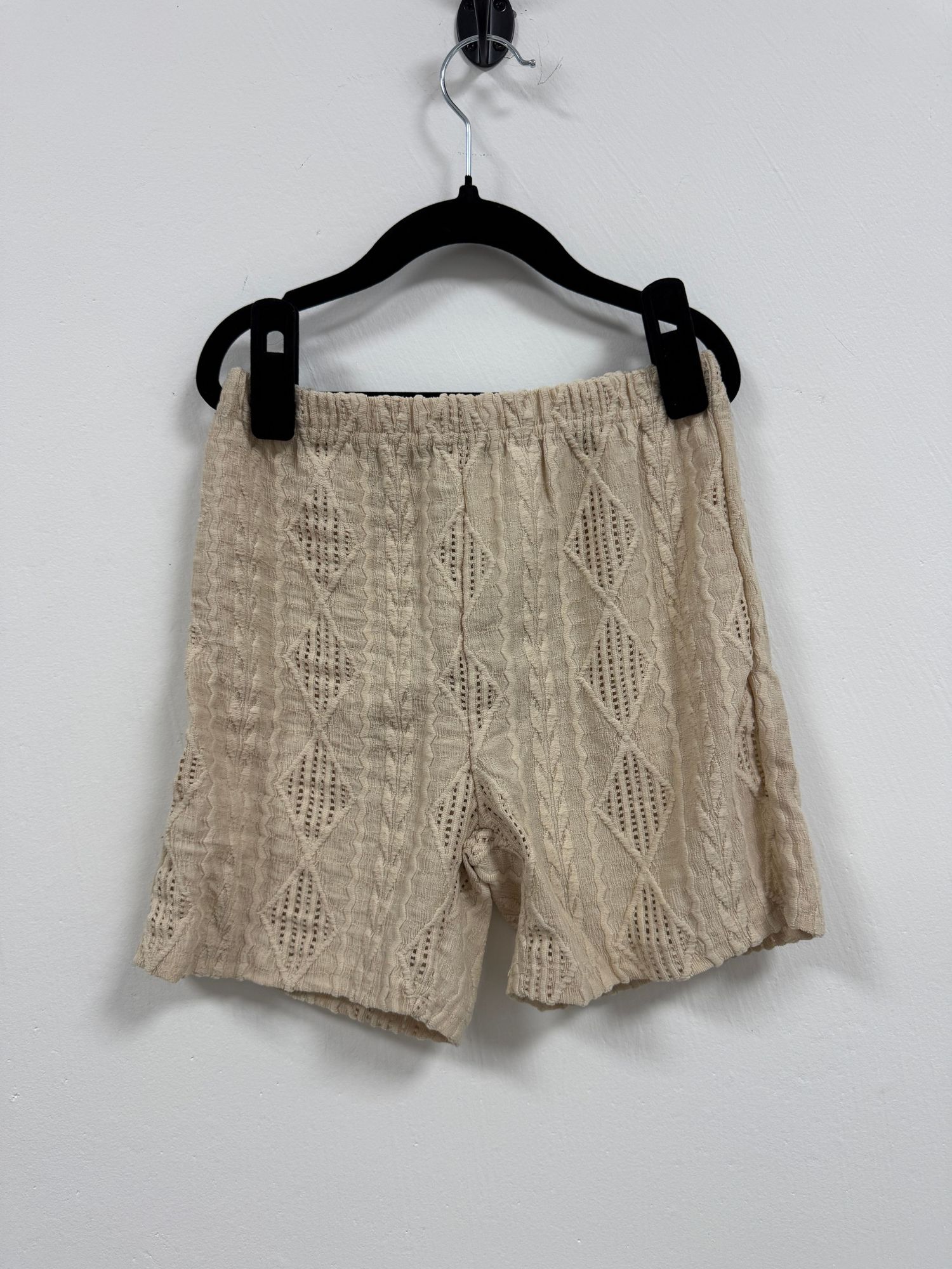 Beige knit textured shorts with diamond pattern hanging on black hanger against white wall.