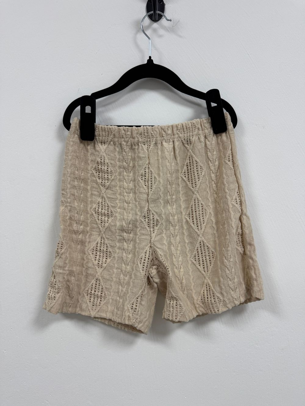 Beige knit textured shorts with diamond pattern hanging on black hanger against white wall.