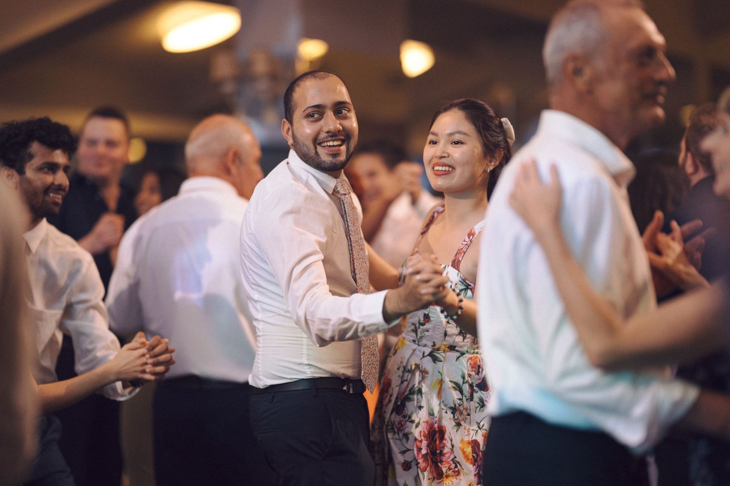 People dancing, laughing and having fun at a wedding reception with warm lighting in the background.