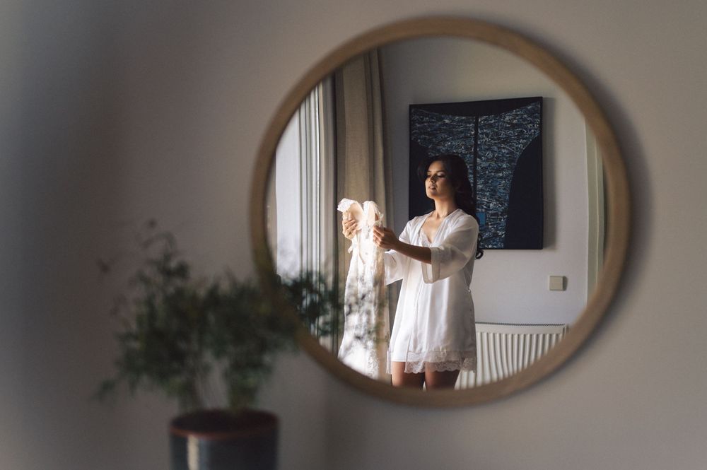 A mirror reflection shows a person standing in white clothing at home, with soft natural light coming through a window.
