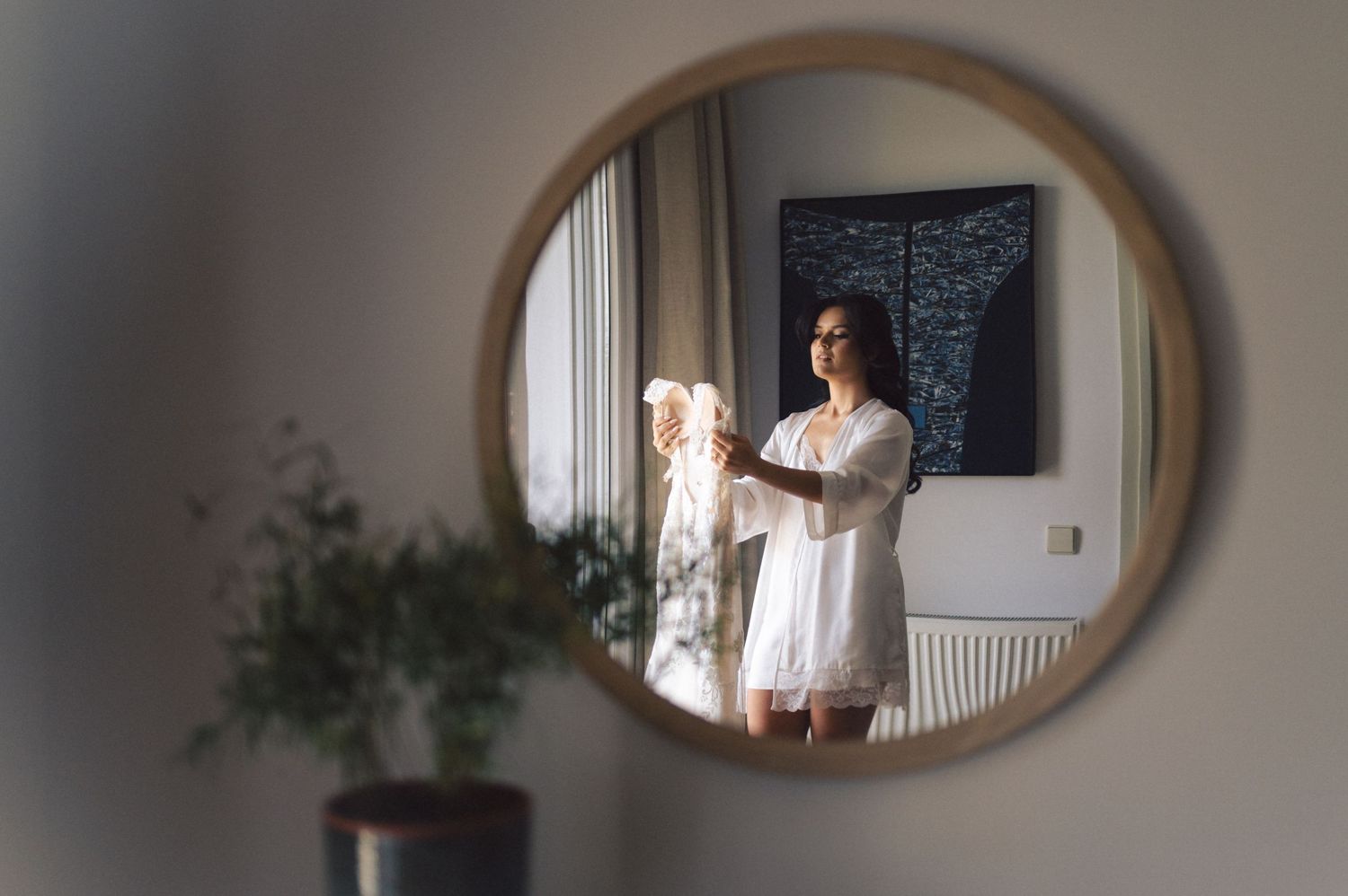 A mirror reflection shows a person standing in white clothing at home, with soft natural light coming through a window.